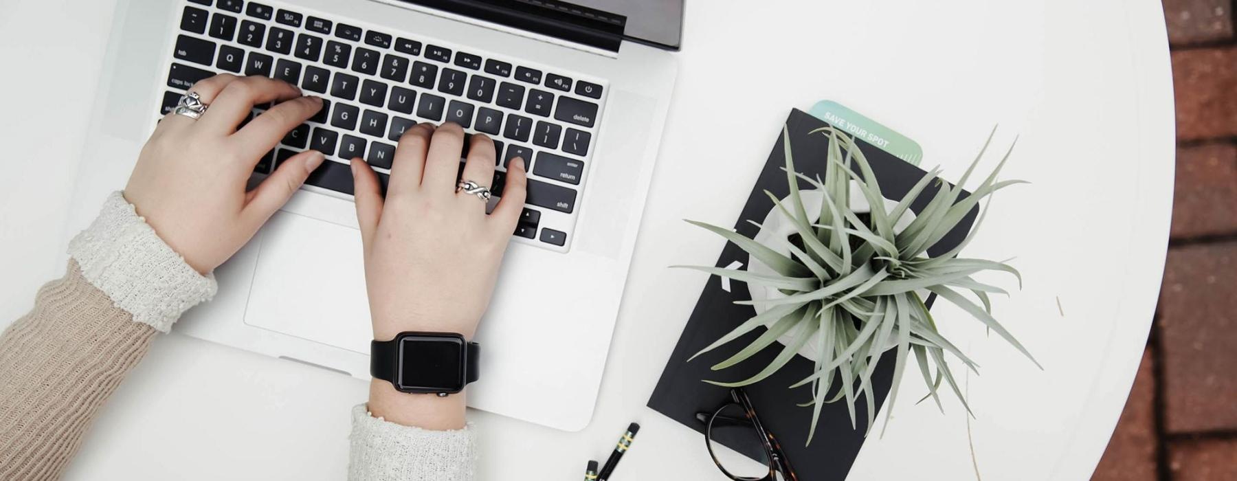 overhead shot of woman working on a laptop at a round table with pencils, her cell phone, glasses, notebook and a potted plant